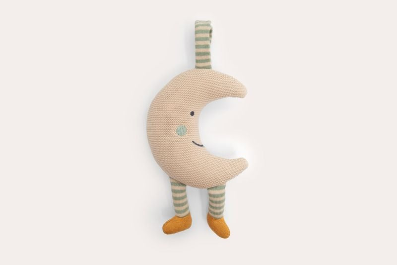 Knitted beige crescent moon with a stitched face, green striped legs and mustard feet, hanging from a knitted tab.