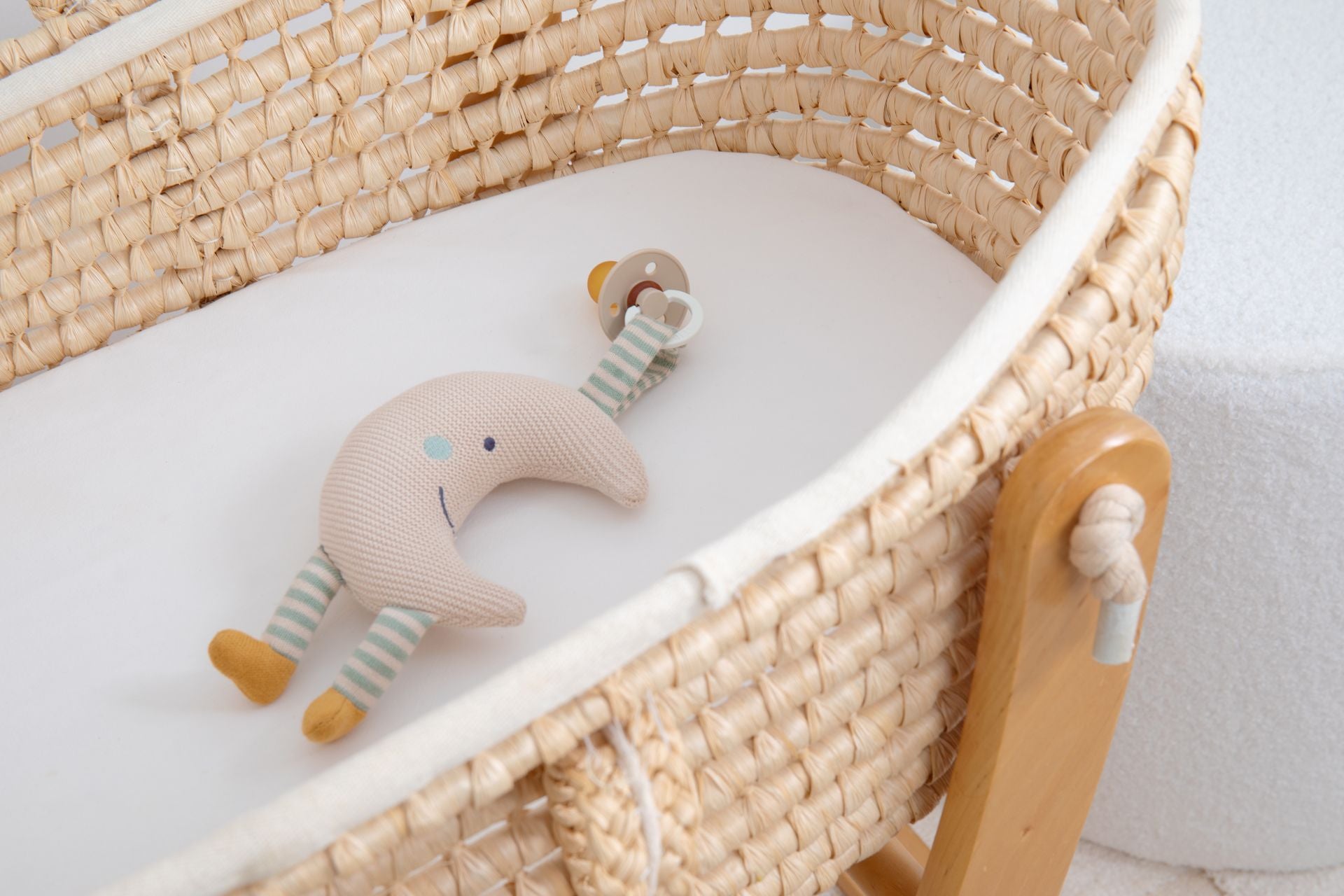 The moon pacifier holder lying in a woven bassinet with a dummy on a wooden ring attached to its tab.