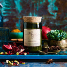 Moroccan Spice candle on a teal setting with pomegranate seeds, a succulent, a brass pot and pistachios