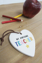 My Awesome Teacher ceramic heart lying on a wooden desk beside an apple and coloured pencils