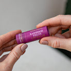 Hands holding the purple-pink Raspberry Fields lip balm tube.
