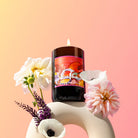 Lit Neon Bloom candle with dahlias in a sculptural vase on a peach backdrop