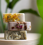 Three handmade soap bars stacked on a pale dish, topped with calendula petals, dried rose and swirled green clay
