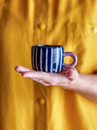 The blue-stripe Nini cup held on an open hand, its lilac handle to the side.