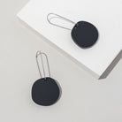Two matte black acrylic discs on long, slim silver-tone ear wires, laid across a white block.