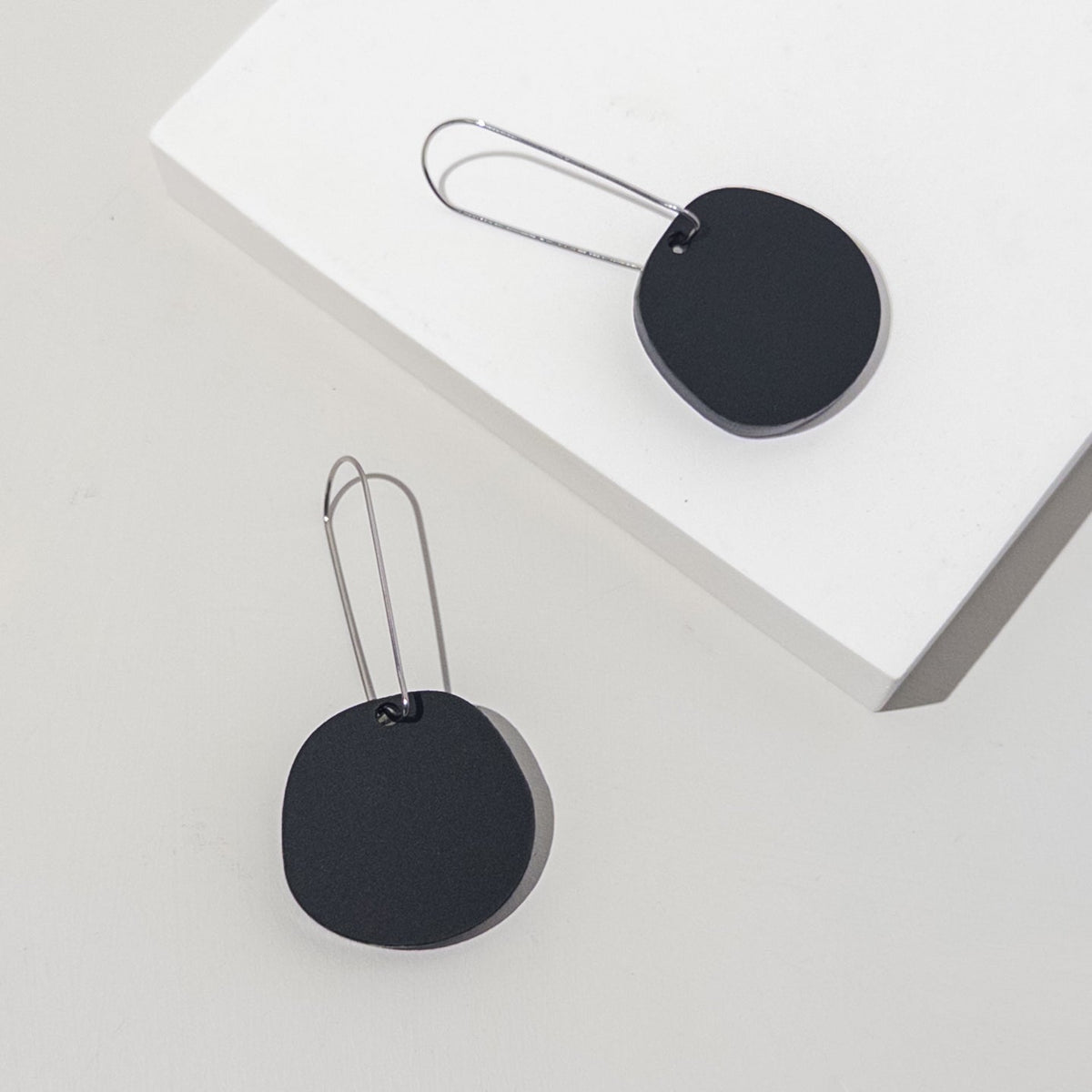 Two matte black acrylic discs on long, slim silver-tone ear wires, laid across a white block.