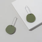 Two olive green acrylic discs on long, slim silver-tone ear wires, laid across a white block.