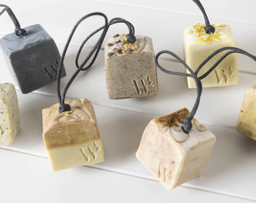 Handmade soaps on rope cords in charcoal, oatmeal, cream and botanical-topped bars, laid on white timber