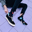 Person crouching on a lilac tiled floor in black sneakers, wearing the black planets and stars socks.