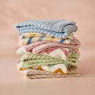 The pink blanket in a folded stack with blue, green, oatmeal, striped and chevron knit blankets from the same range, on a peach background