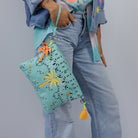 The aqua palm tree pouch carried by its wrist strap against light denim jeans.