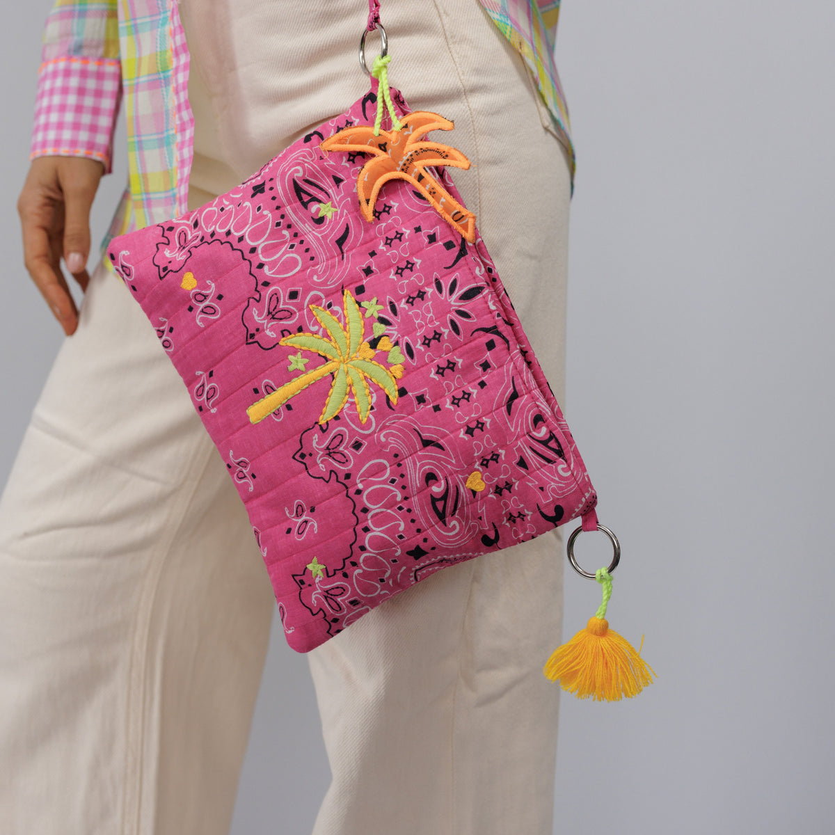 The pink palm tree pouch held by its wrist strap against cream trousers.