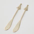 Two gold-tone cheese spreaders side by side on white, with rounded blades and handles topped by a ridged Christmas tree and star