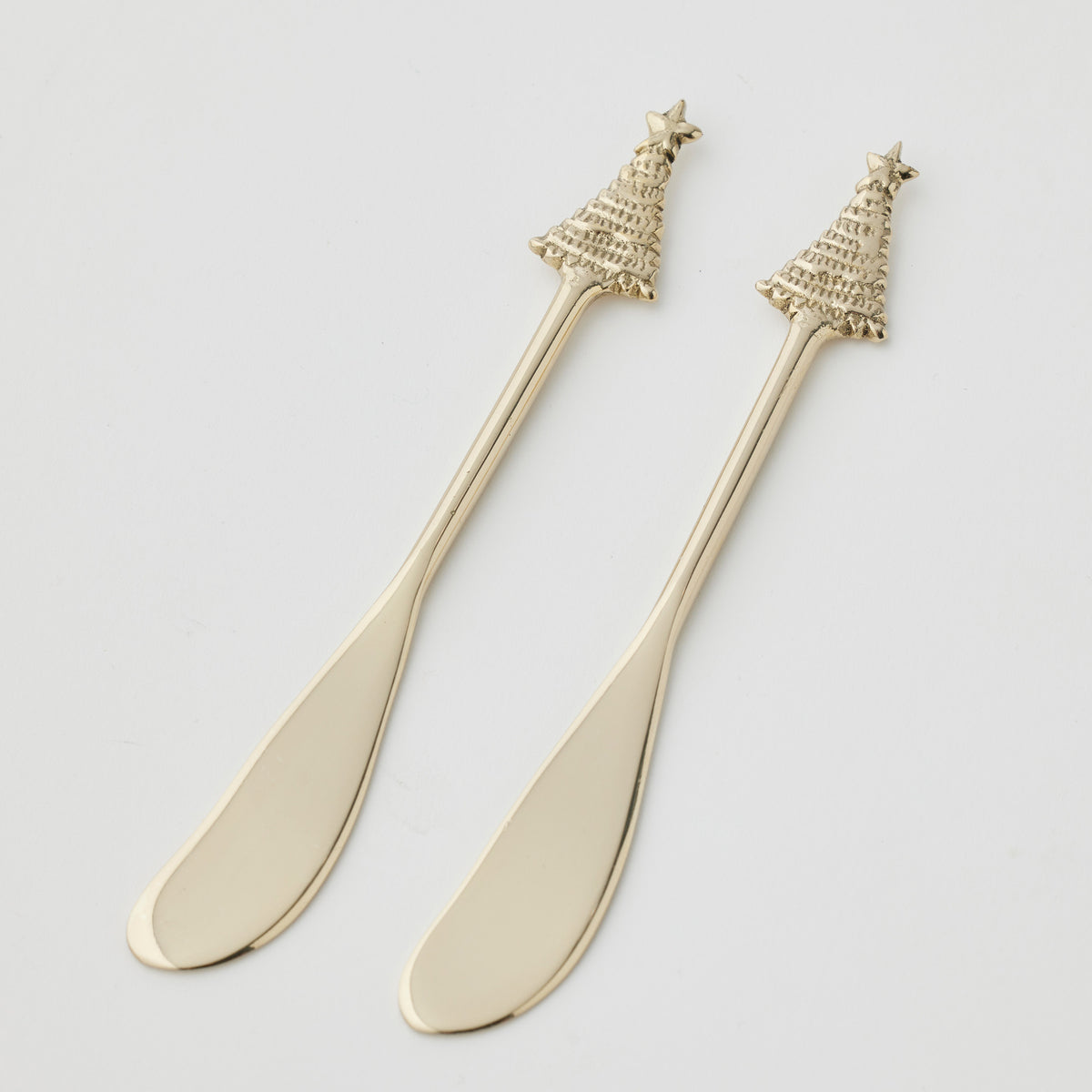 Two gold-tone cheese spreaders side by side on white, with rounded blades and handles topped by a ridged Christmas tree and star