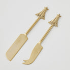 Two gold-tone cheese knives on white, one with a wide flat blade and one with a curved forked tip, both with Christmas tree and star handles