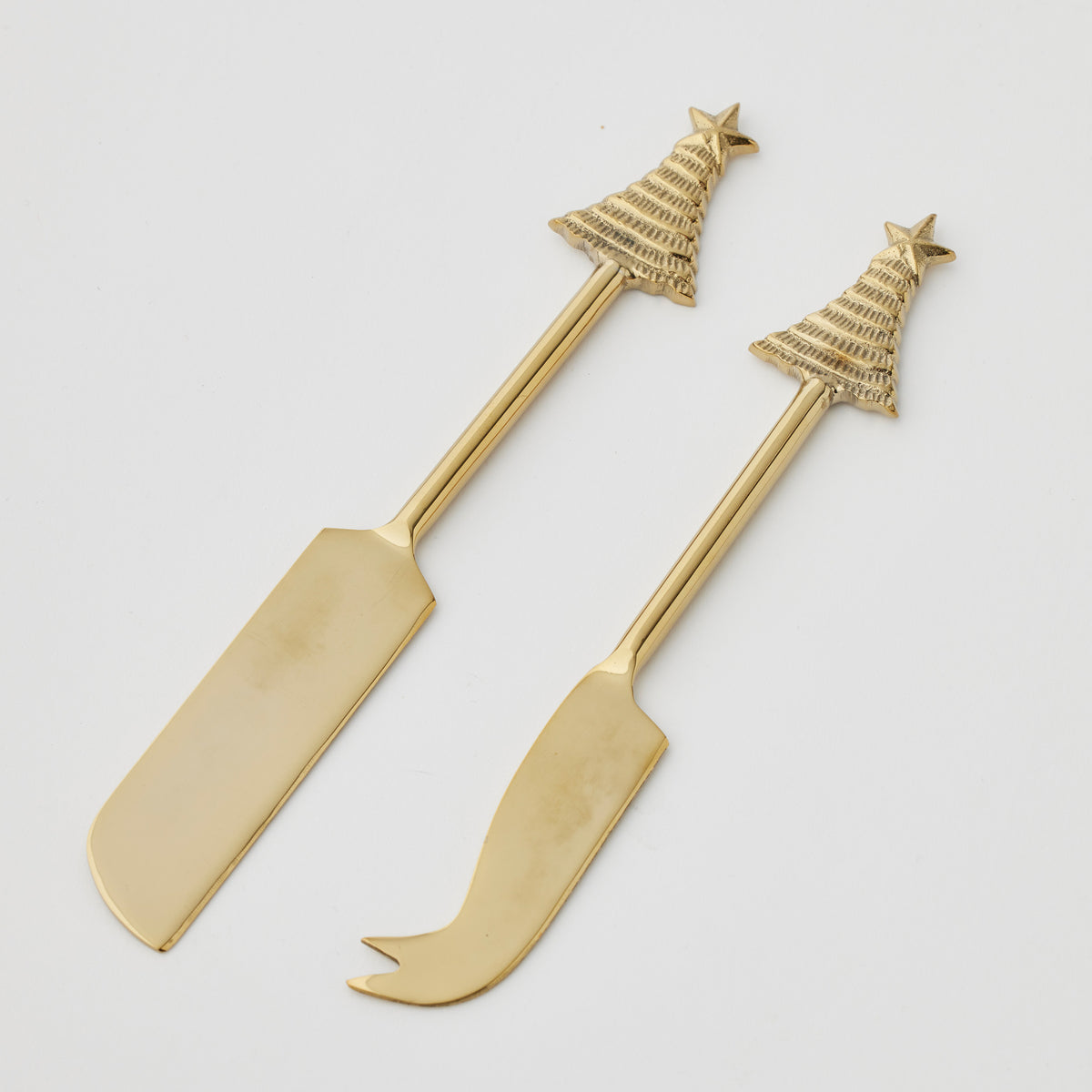 Two gold-tone cheese knives on white, one with a wide flat blade and one with a curved forked tip, both with Christmas tree and star handles