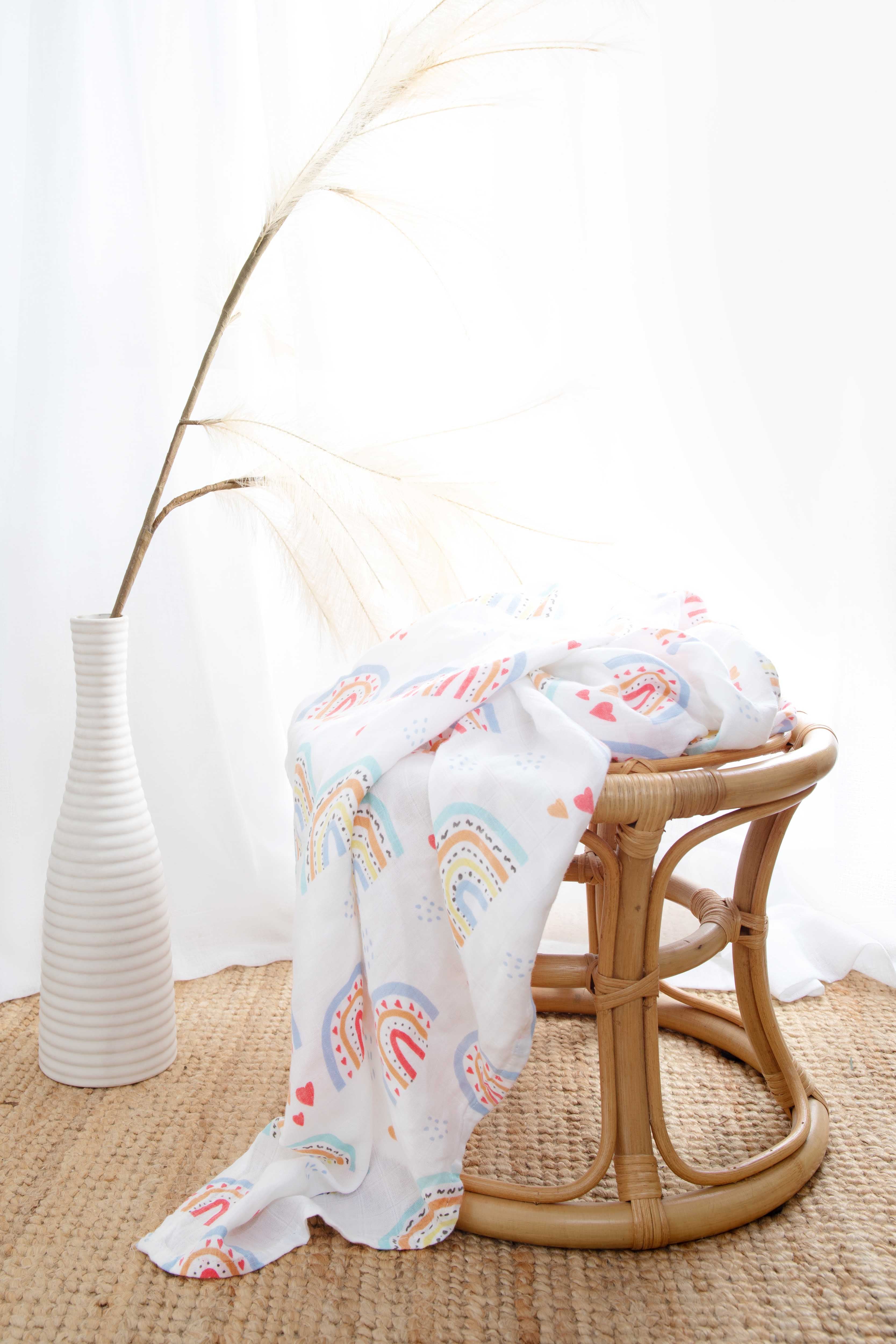 The rainbow-print muslin wrap draped over a rattan stool beside a white vase of pampas grass.