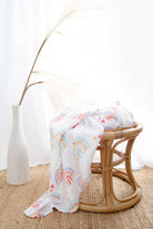 The rainbow-print muslin wrap draped over a rattan stool beside a white vase of pampas grass.