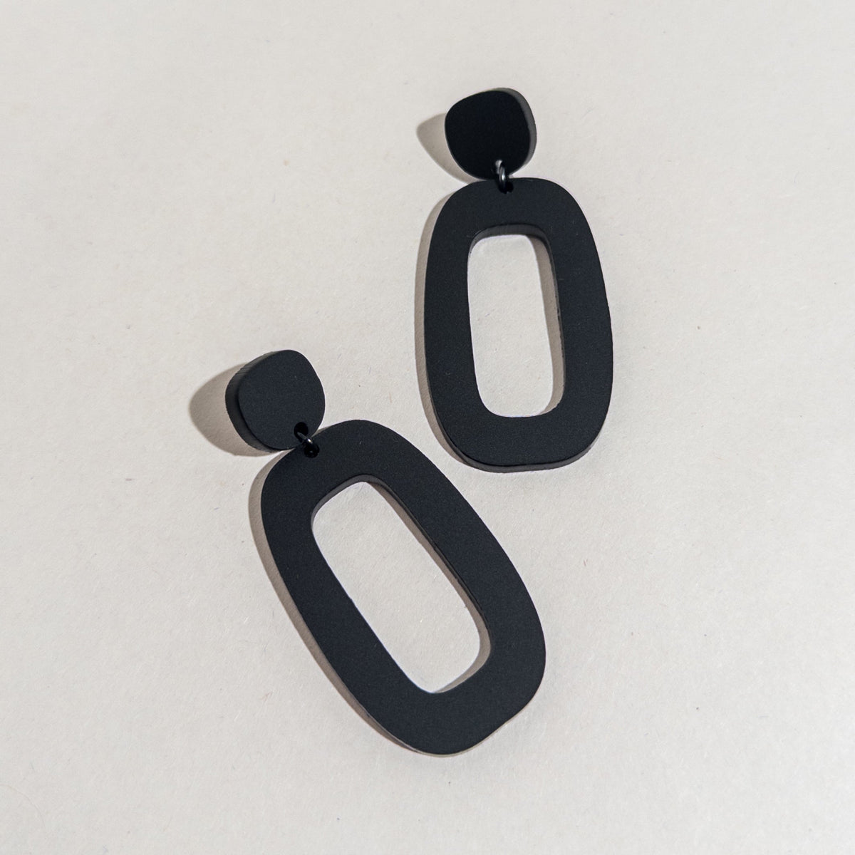 A pair of matte black acrylic dangle earrings on a white surface, each an open oval hung from a small rounded top.