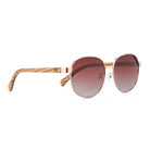 Cleo Bloom sunglasses at an angle: a round rose gold metal frame, brown gradient lenses and striped walnut arms