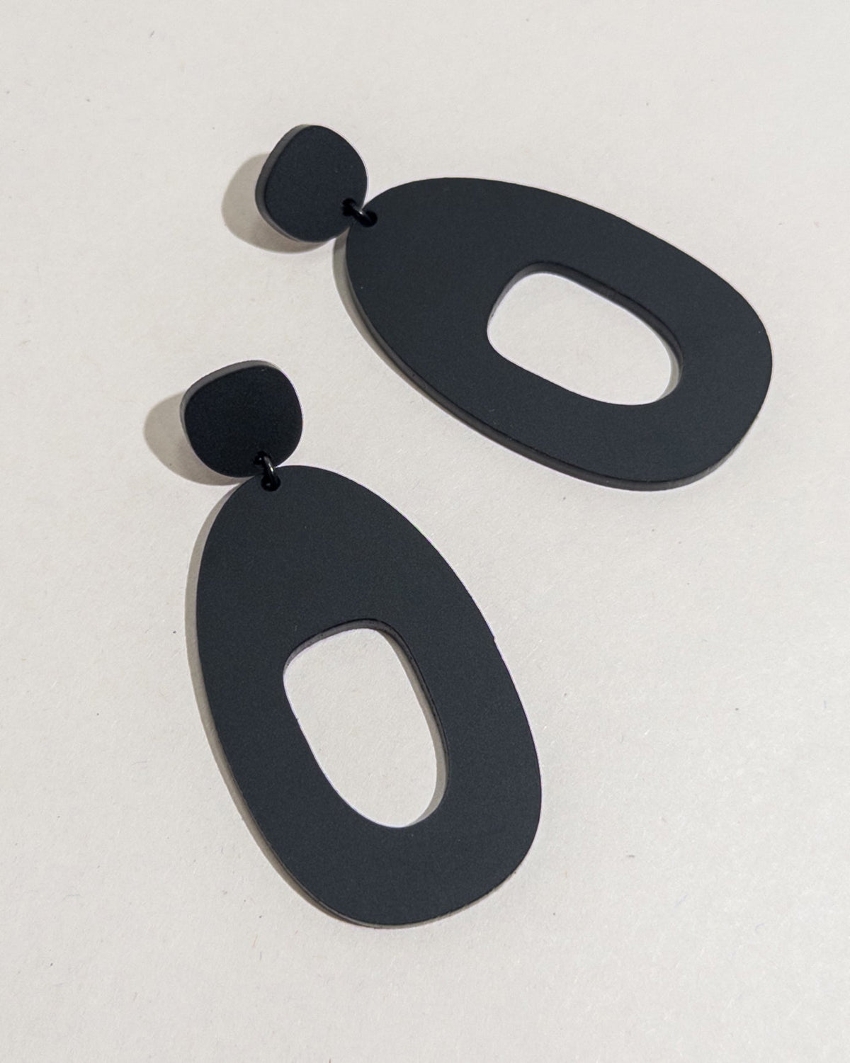 A pair of large matte black acrylic statement earrings, each a wide open shape hung from a small rounded top.