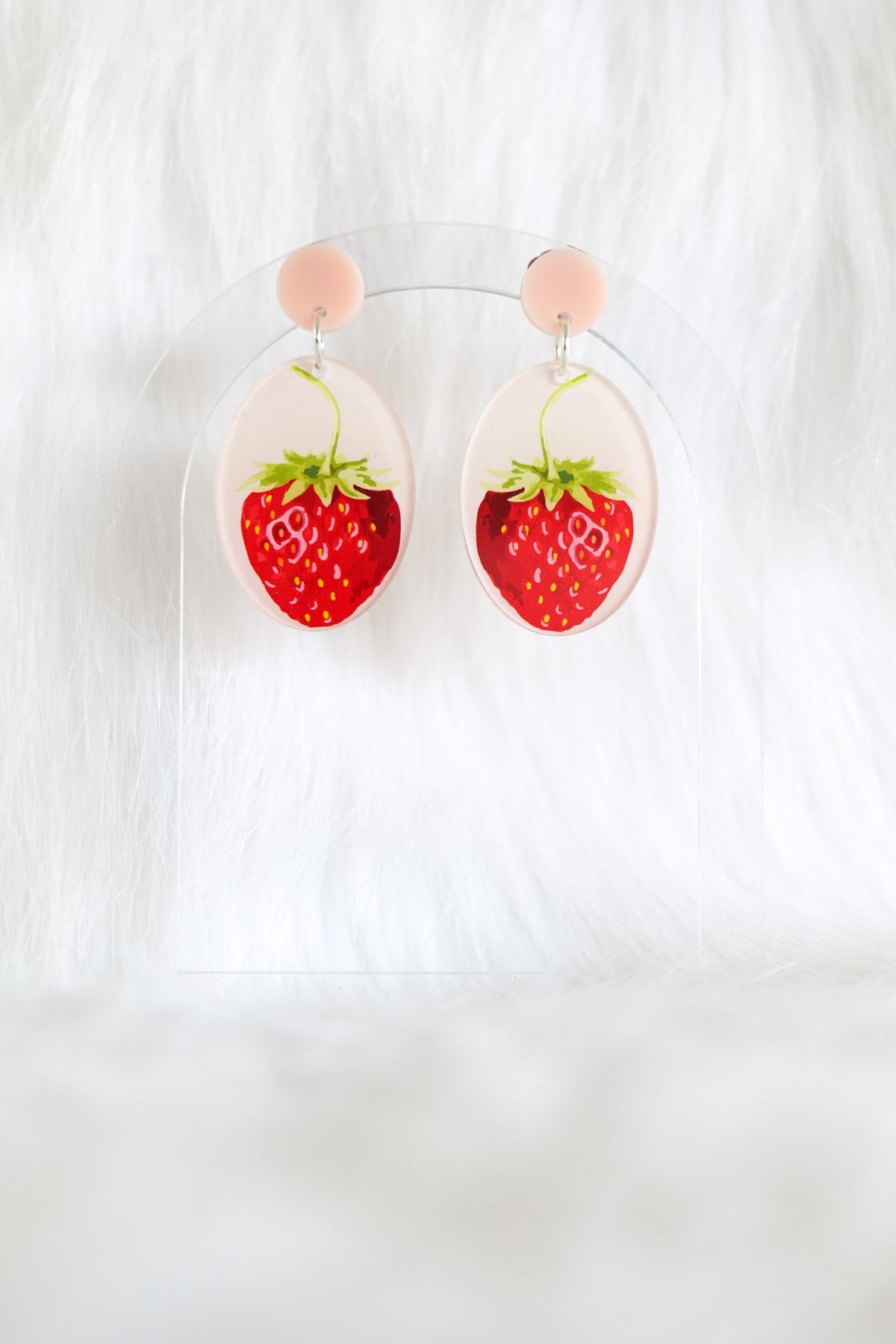 The strawberry earrings hanging on a clear stand: pale pink studs above clear ovals with red strawberries.