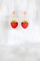 The strawberry earrings hanging on a clear stand: pale pink studs above clear ovals with red strawberries.