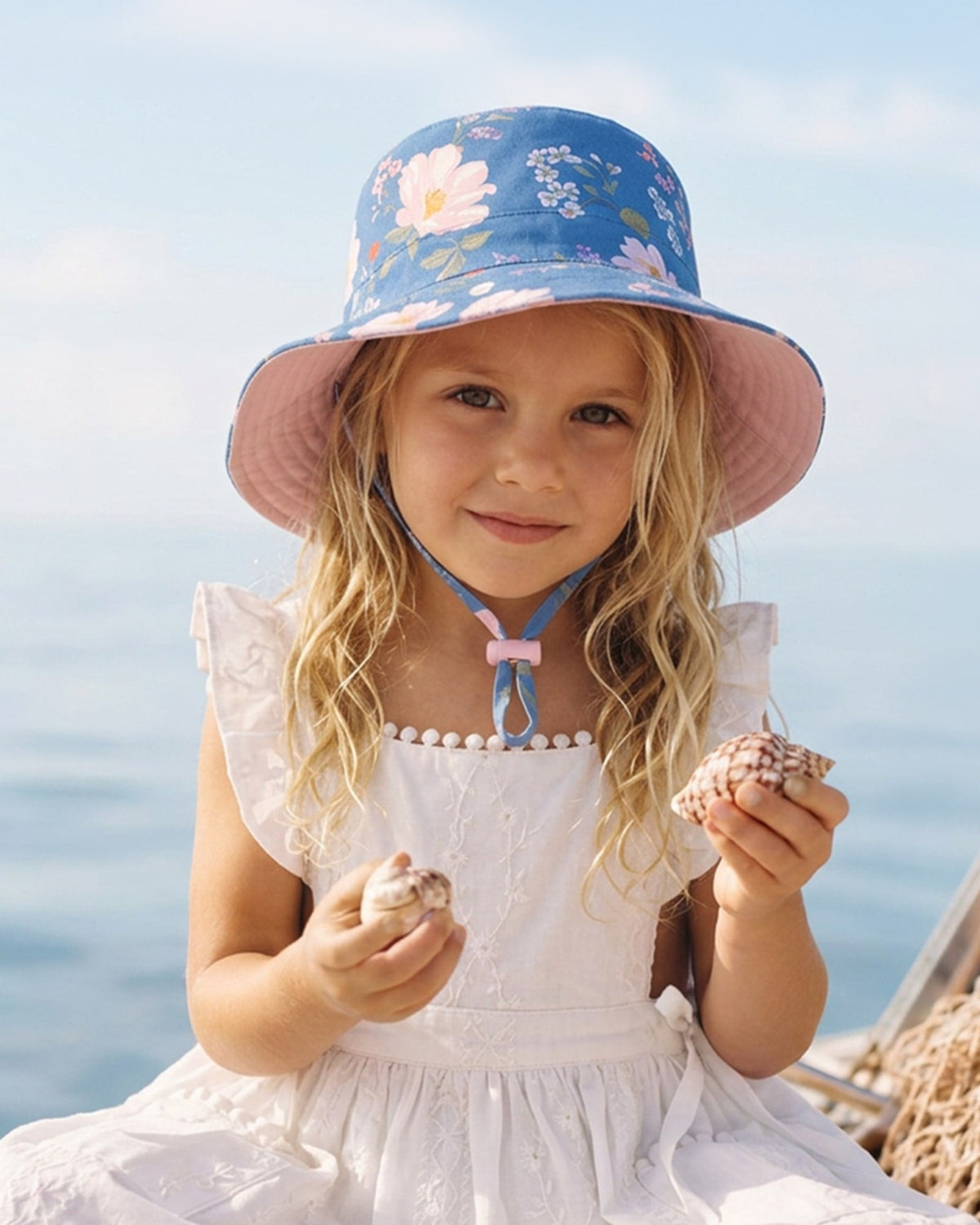 Girl by the sea in the Camille sunhat and a white frilled dress, holding shells