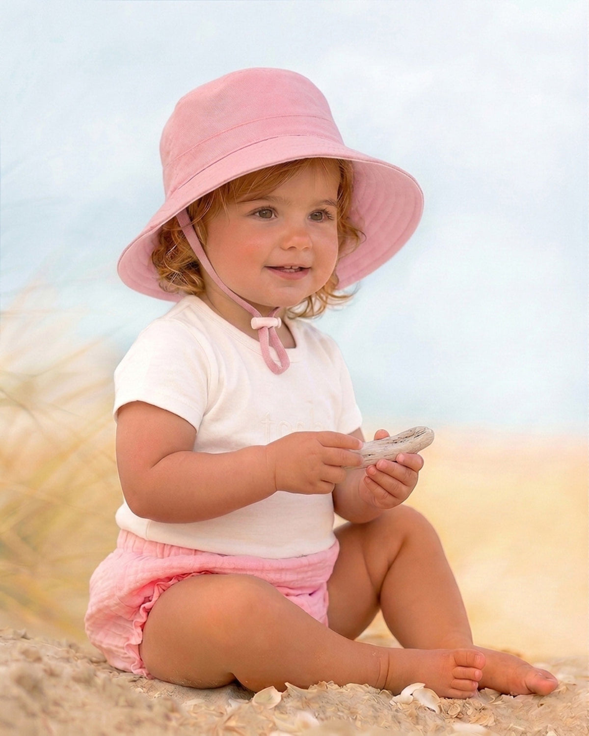 Toddler sitting on the sand in the pink Olly sunhat