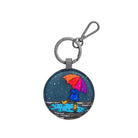 Round Rainy Day keyring: a figure in a blue raincoat and yellow boots under an orange, pink and purple umbrella, splashing in a puddle on slate blue.