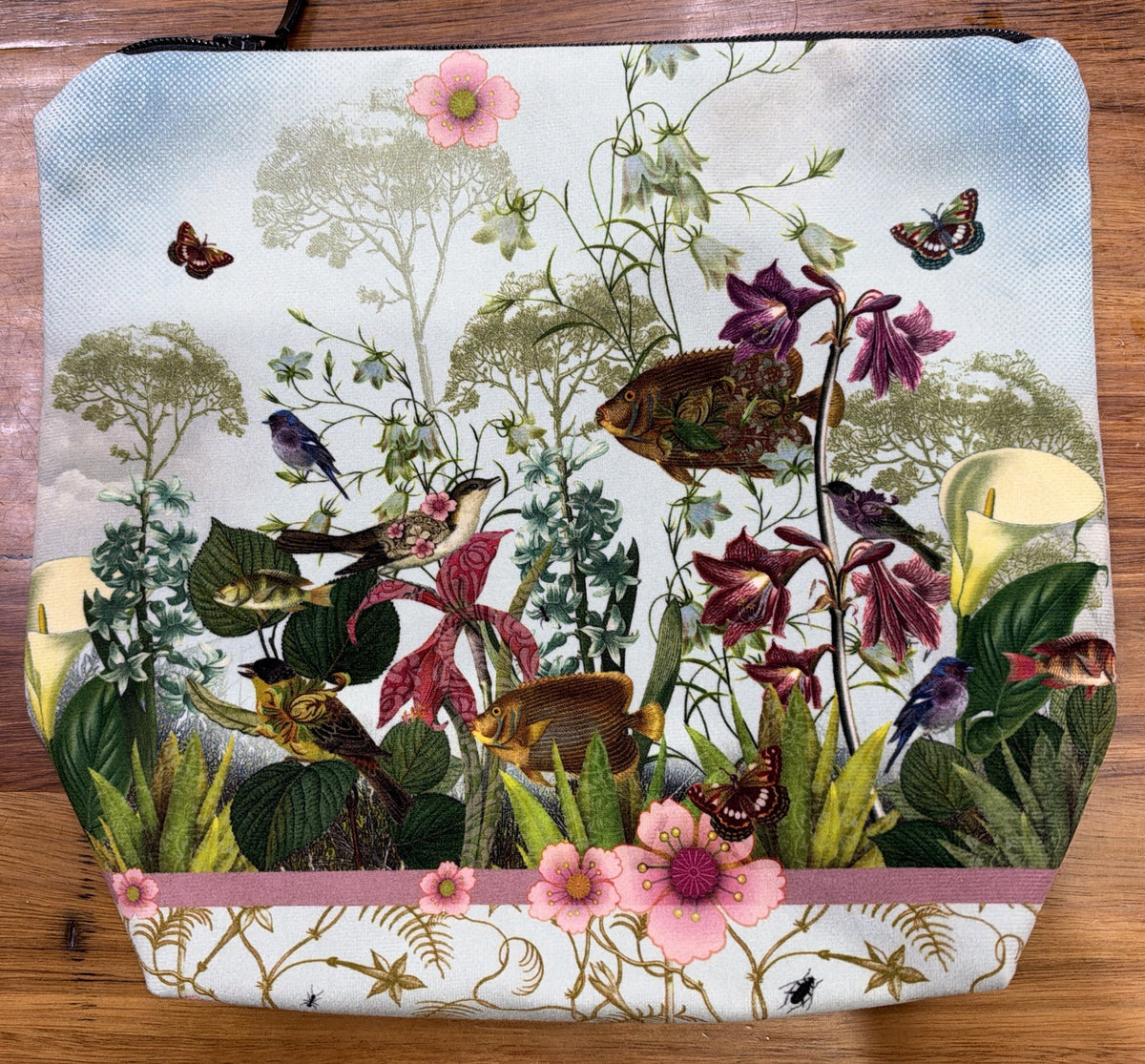 Velour cosmetic bag printed with Isabelle Frioux's Happiness in the Meadow: birds, fish and butterflies among pink flowers under a pale blue sky, with a black zip.