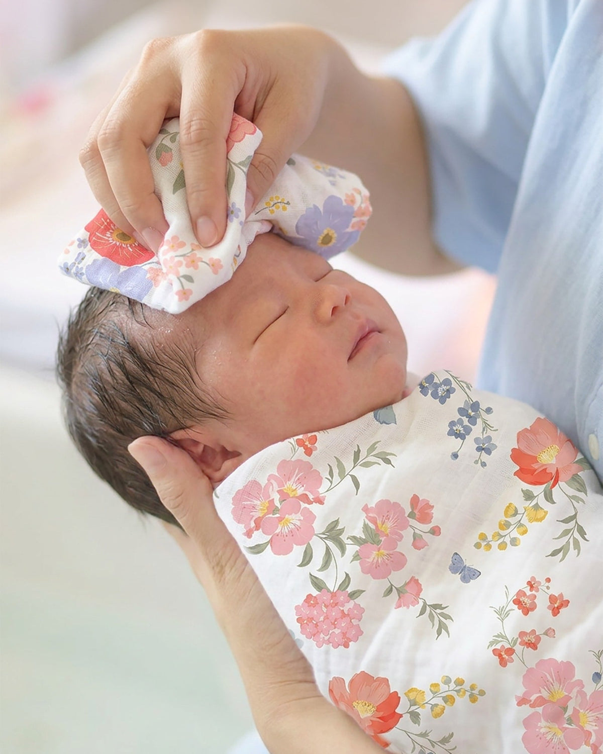 Newborn swaddled in the Wild Garden muslin wrap having their forehead dabbed with a coordinating floral washcloth
