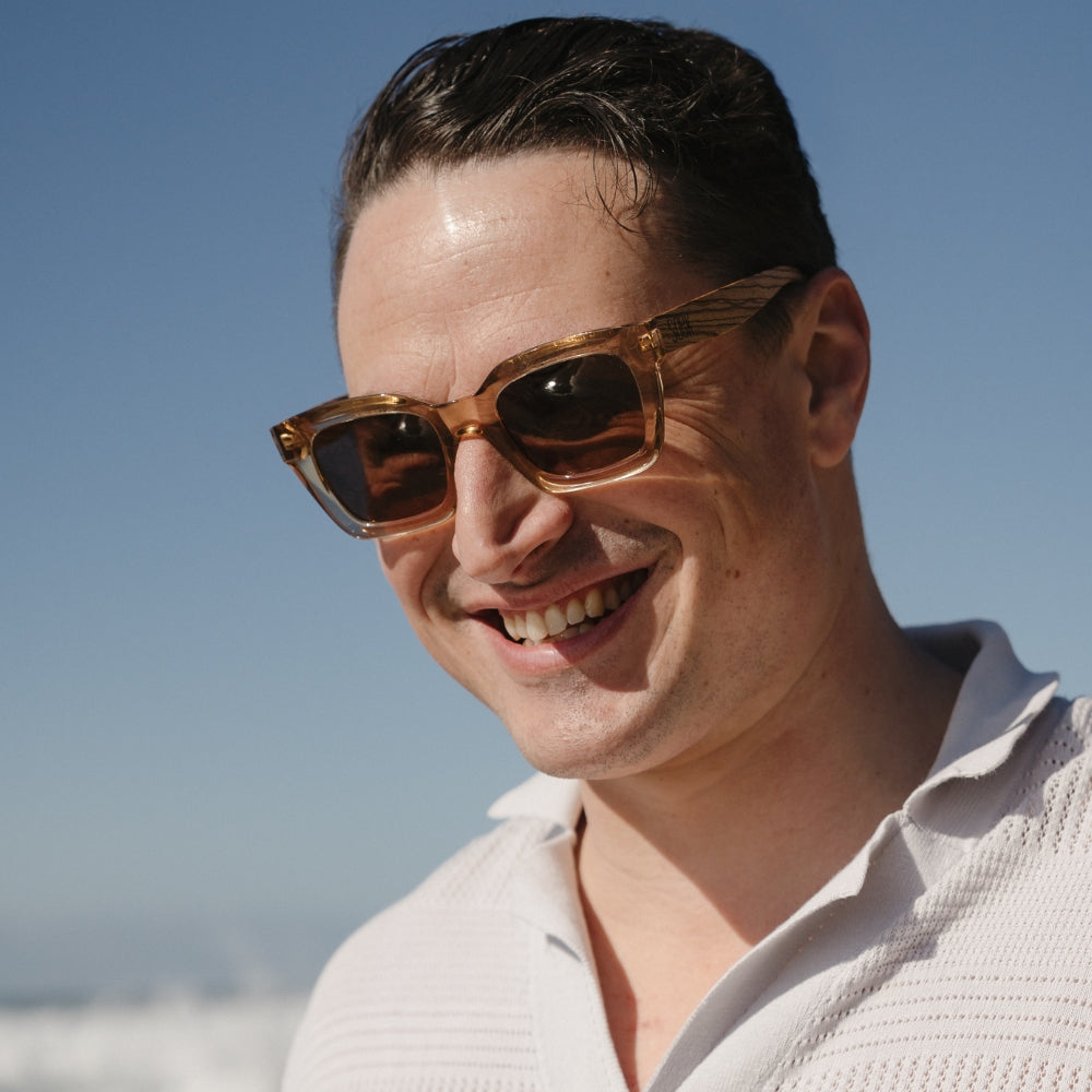 A smiling man with dark hair wearing the Zahra Champagne sunglasses against a blue sky