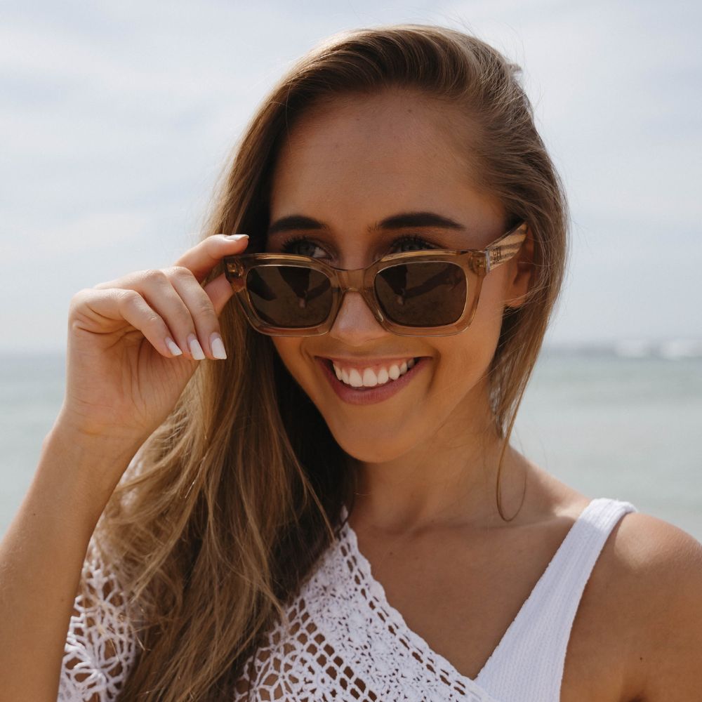 A smiling woman lifting the Zahra Champagne sunglasses with one hand, the sea behind her