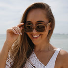 A smiling woman lifting the Zahra Champagne sunglasses with one hand, the sea behind her