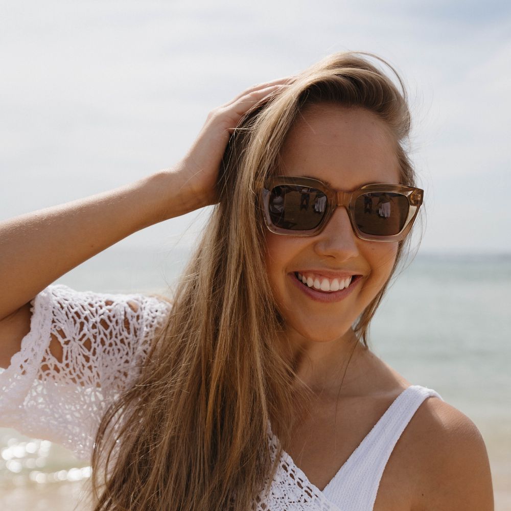A woman with long brown hair smiling at the beach in the Zahra Champagne sunglasses, one hand in her hair