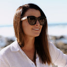 A woman with long brown hair looking off to the side in the Milla Khaki sunglasses, the sea behind her